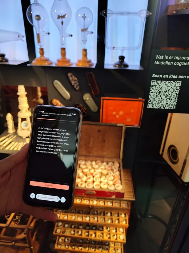 Bezoeker houdt een telefoon voor de vitrine, met op het scherm een verhaal over glazen oogmodellen